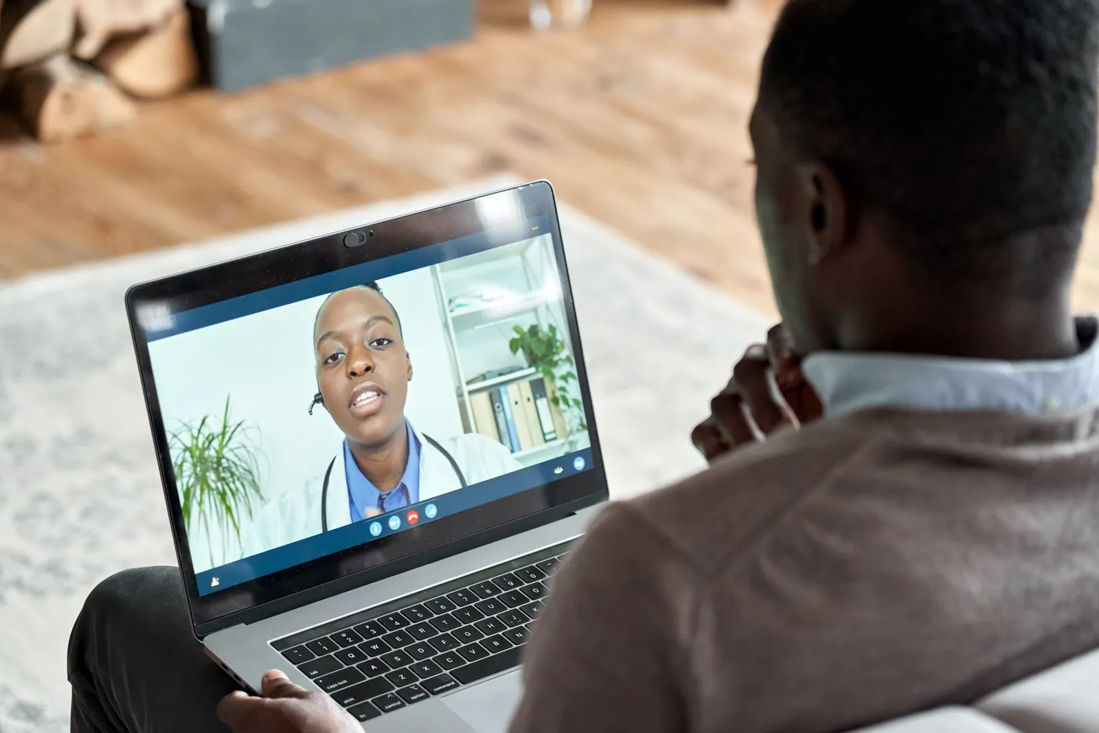 Physician video-calling a patient from a laptop during a virtual telehealth visit, stethoscope and notepad on the desk beside the screen