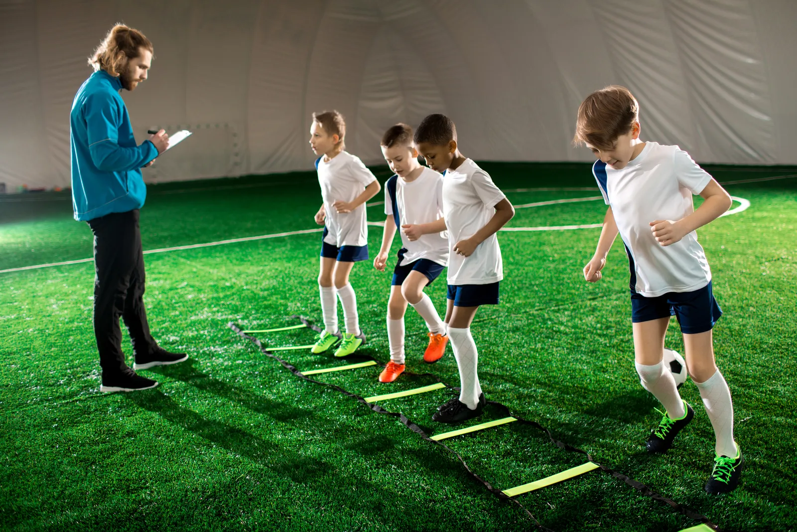 Youth sports coach guiding a small group of kids through an agility ladder drill on an indoor turf field