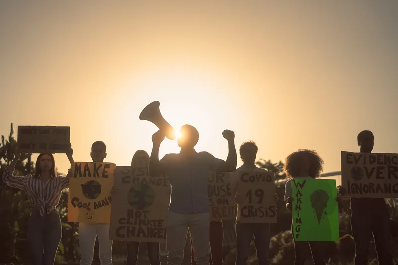 Climate marchers silhouetted against a sunset sky, one raising a megaphone, holding hand-lettered signs