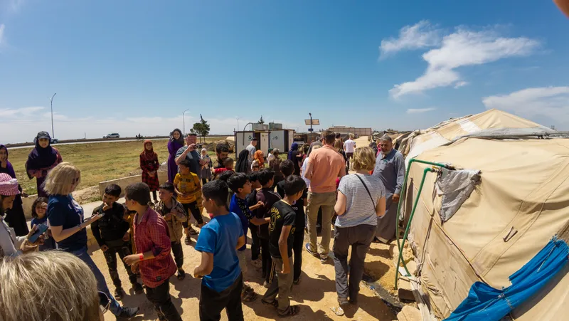 Families and children gathered on a sunlit path beside a row of canvas shelters, talking with visiting aid workers