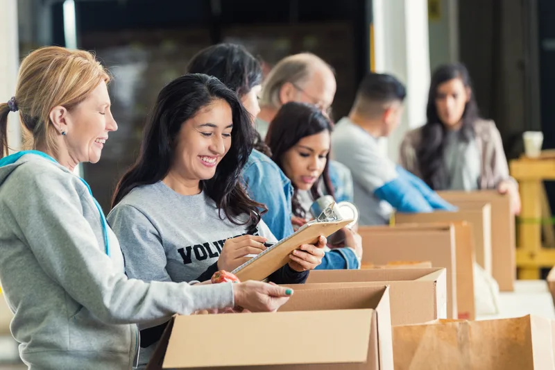 Volunteers in Riverbend sweatshirts laughing together while packing grocery boxes, one checking a route list on a clipboard