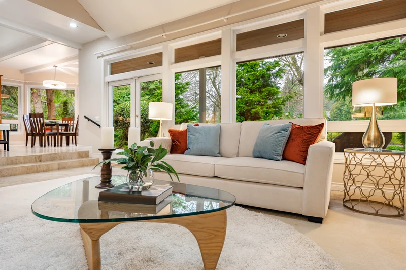 Sunlit living room with a cream sofa, wall of windows onto mature trees and steps up to the dining area