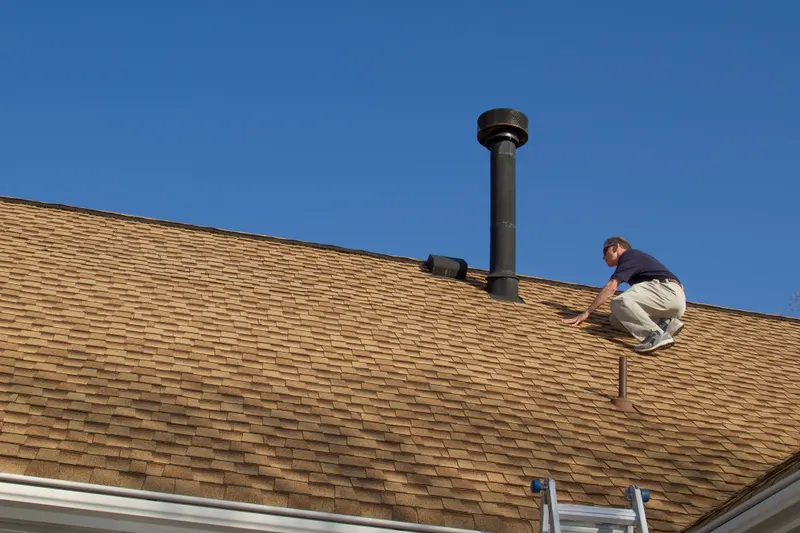 Inspector crouched on an asphalt shingle roof examining the flue and vent flashing