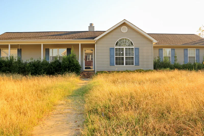 A vacant ranch house with blue shutters behind a front yard of tall, dry, overgrown grass