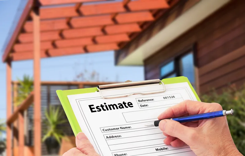 A hand filling in a repair estimate form on a clipboard outside a house