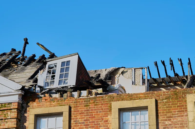 The burned-out roof and dormer of a brick house after a fire, against a clear blue sky