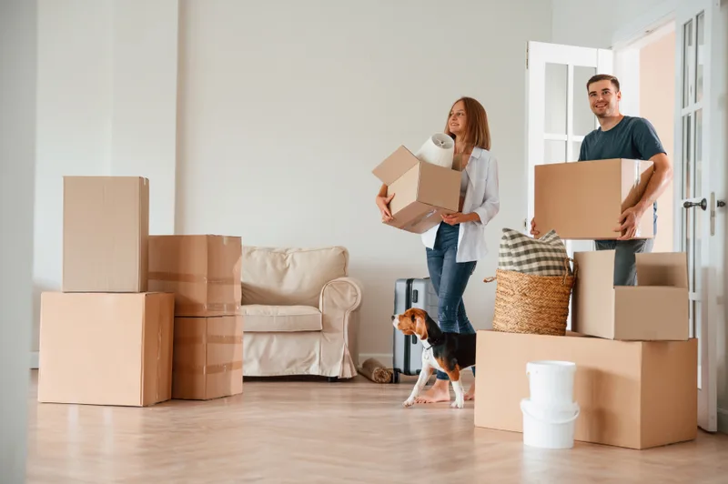 A couple carrying moving boxes into a bright living room while their dog watches