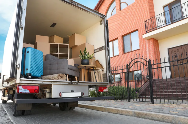 A moving truck parked outside a brick home, loaded with boxes, furniture and a blue suitcase