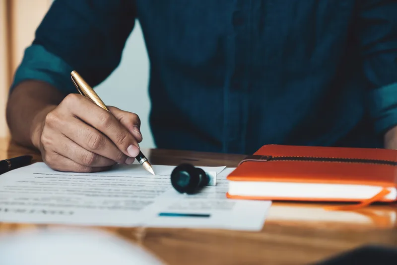 A homeowner's hand writing on mortgage paperwork at a wooden table, next to an orange notebook