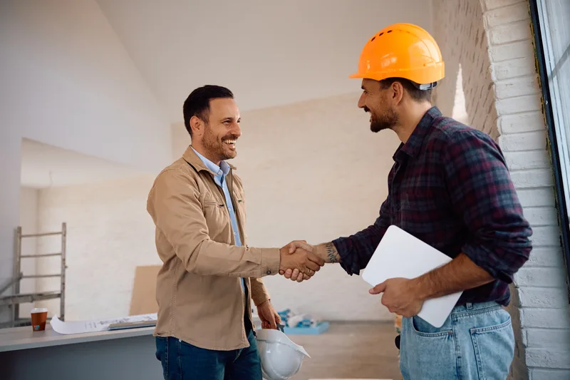 A homeowner and a contractor in a hard hat shaking hands inside a house under renovation