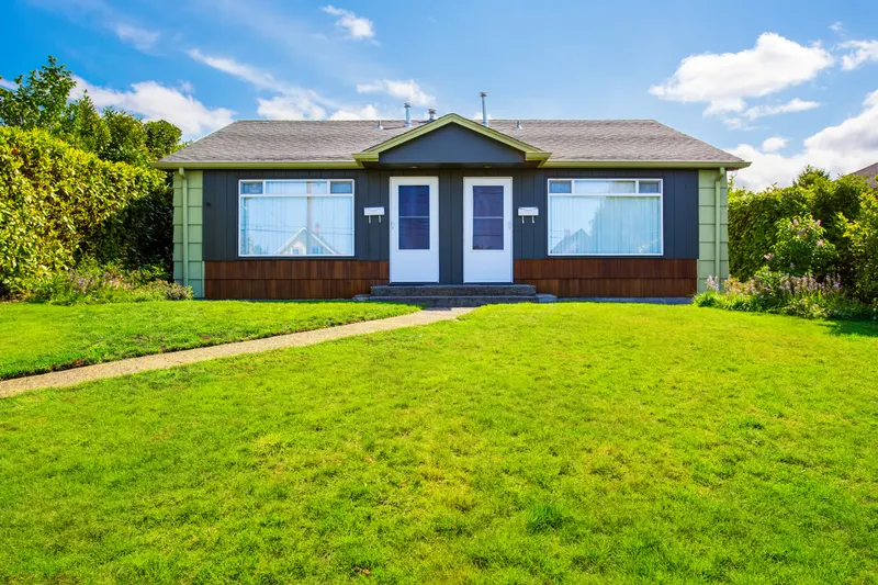 Single-storey two-family duplex with dark siding, twin white front doors and a shared concrete walk across a wide lawn