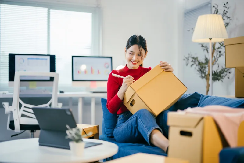 Woman on a sofa writing a label on a cardboard moving box