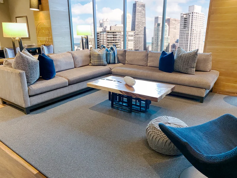 A composed sitting room high above the city, indigo cushions and a live-edge table before a wall of tower glass