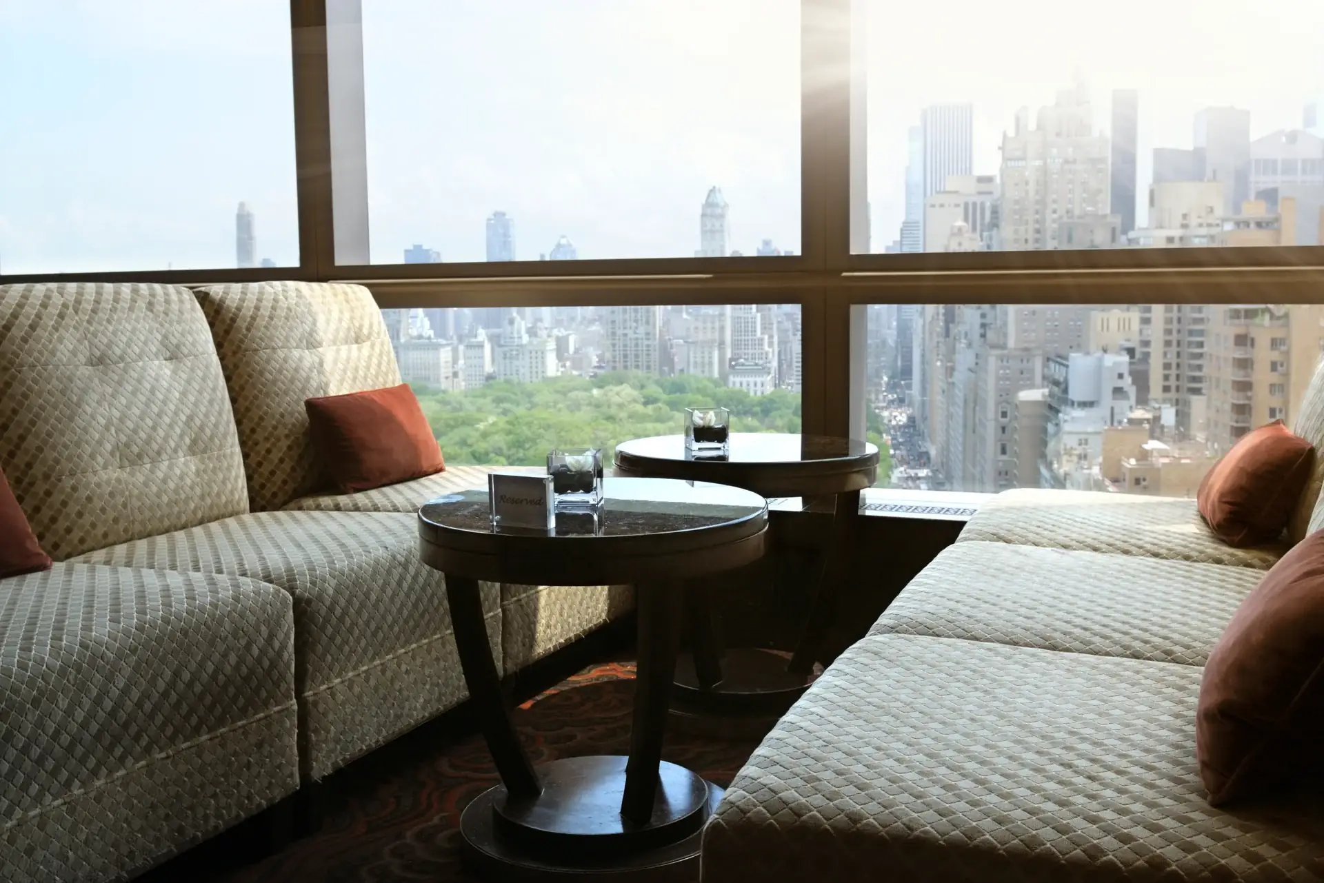 Residence 27's window lounge above Alder Park — quilted ivory sofas, low mahogany tables, and the green canopy stretching to the towers beyond