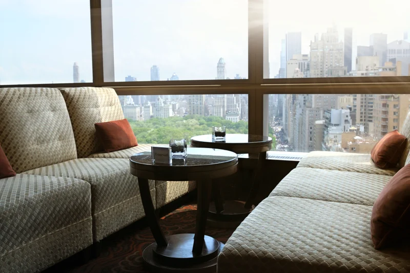 Residence 27's window lounge above Alder Park — quilted ivory sofas, low mahogany tables, and the green canopy stretching to the towers beyond