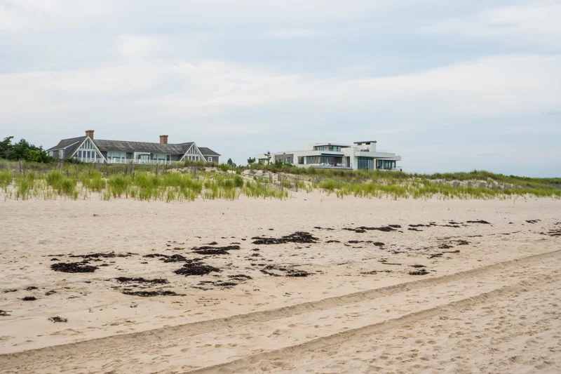 The Shingle House's twin-gabled silhouette above the dune grass, its modern white neighbor visible down the empty beach