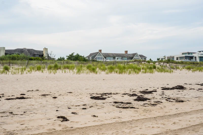 The Saltcliff dune line at midday — shingle rooftops and one white modern volume above the seagrass, tide marks on the empty sand