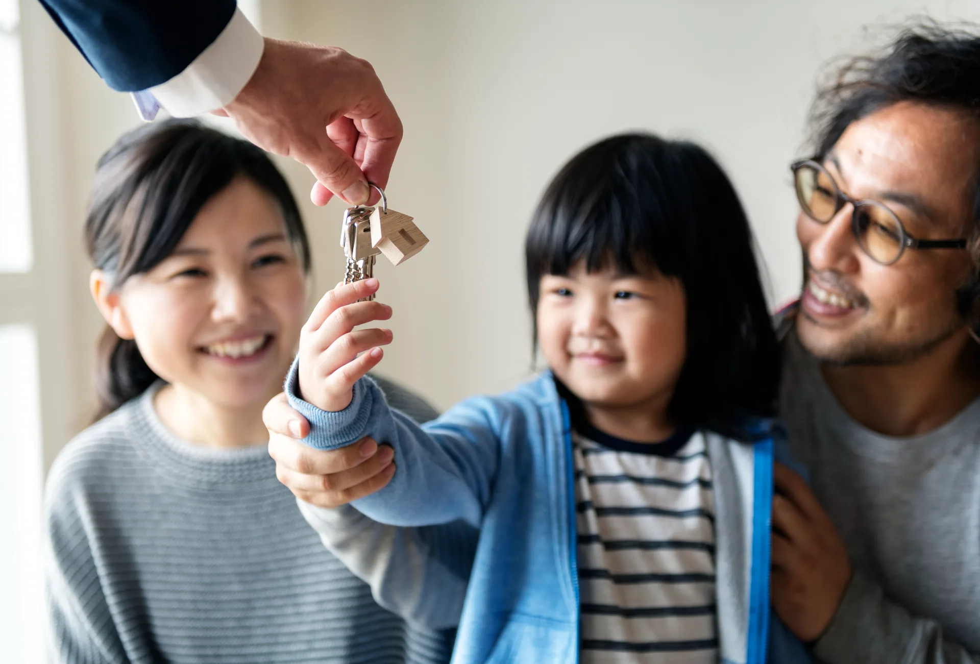 A young child reaching up to take the house keys from the agent's hand, parents smiling on either side