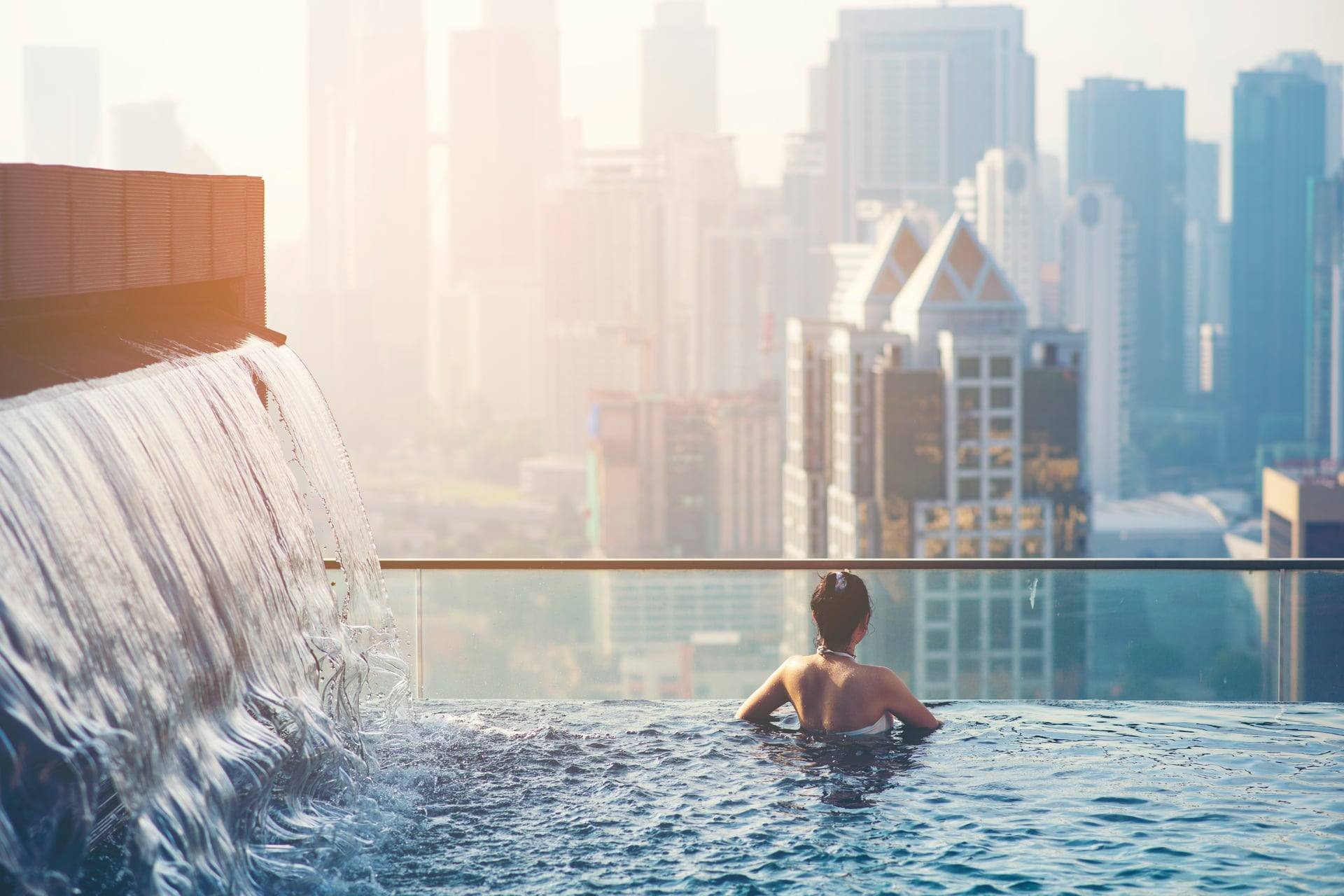 Resident at the infinity edge of a rooftop pool with a cascade water wall, looking out over a sunlit high-rise skyline — artist's impression.