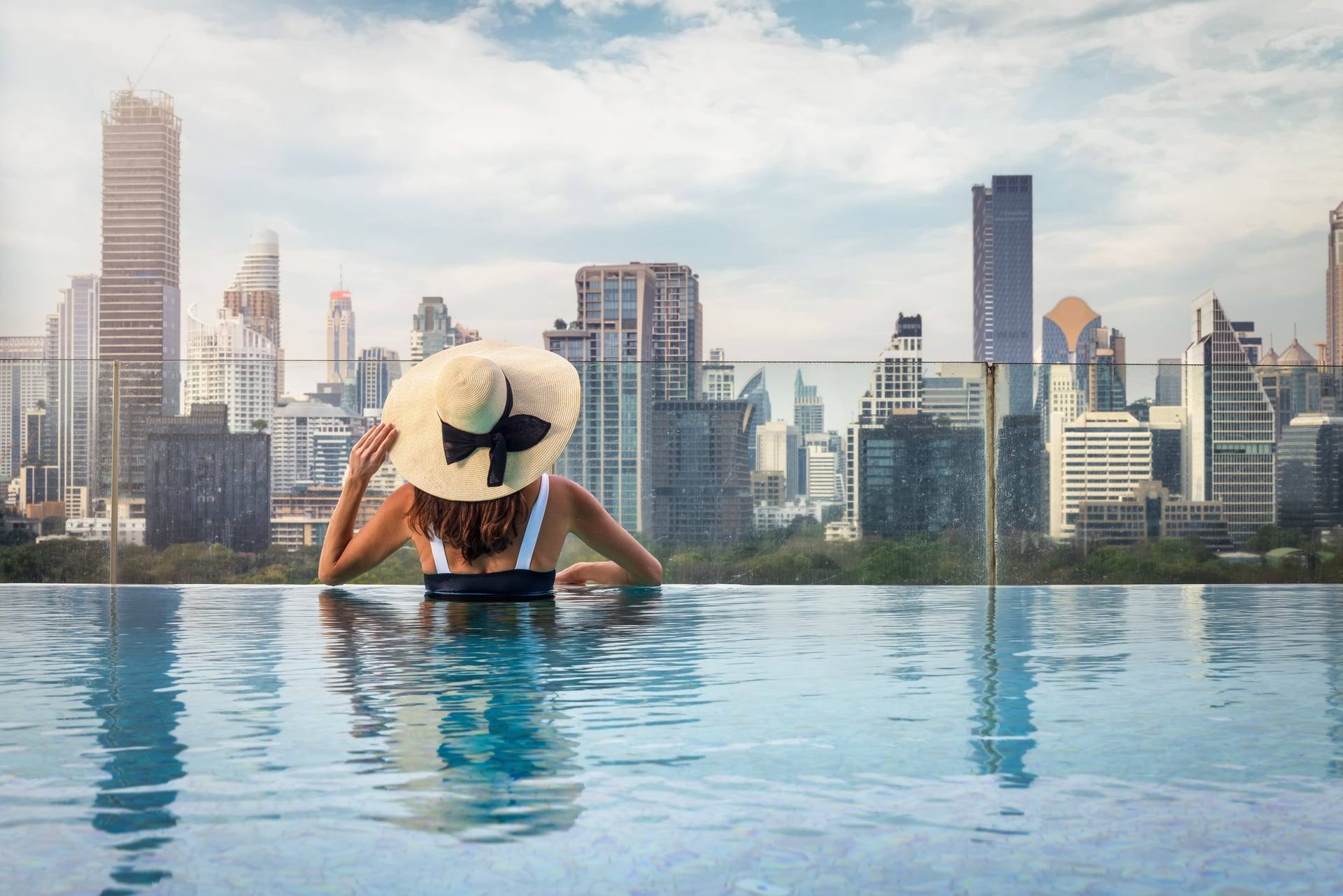 Resident in a sun hat at the edge of an infinity pool, the city skyline spread beyond the glass rail — artist's impression.