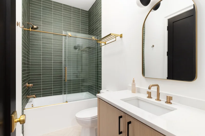 Renovated bathroom with green stacked tile, a glass shower door over the tub, brass fixtures and an arched mirror
