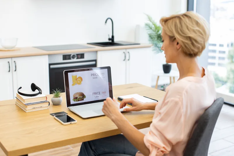 Woman at a kitchen table ordering lunch from a laptop, phone beside her