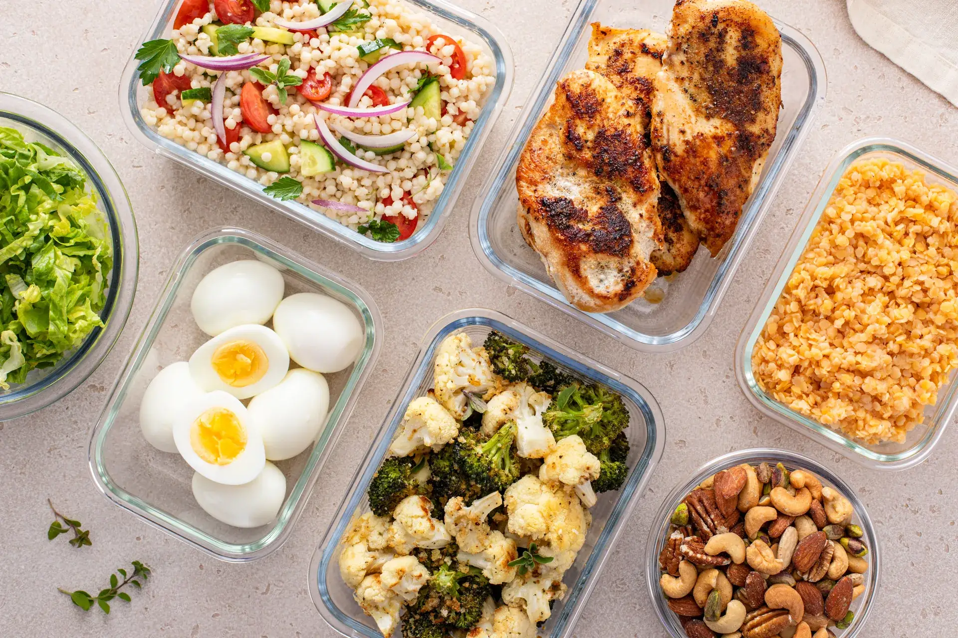 Catering spread of glass containers with grilled chicken, couscous salad, roasted broccoli and cauliflower, boiled eggs, red lentils, and mixed nuts
