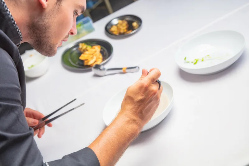 A Brassica cook leaning over the pass, finishing plates of the tasting menu on a white counter lined with small dark dishes