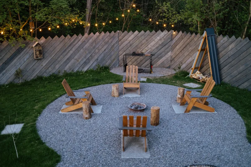 A gravel fire pit circle with wooden lawn chairs under string lights at dusk