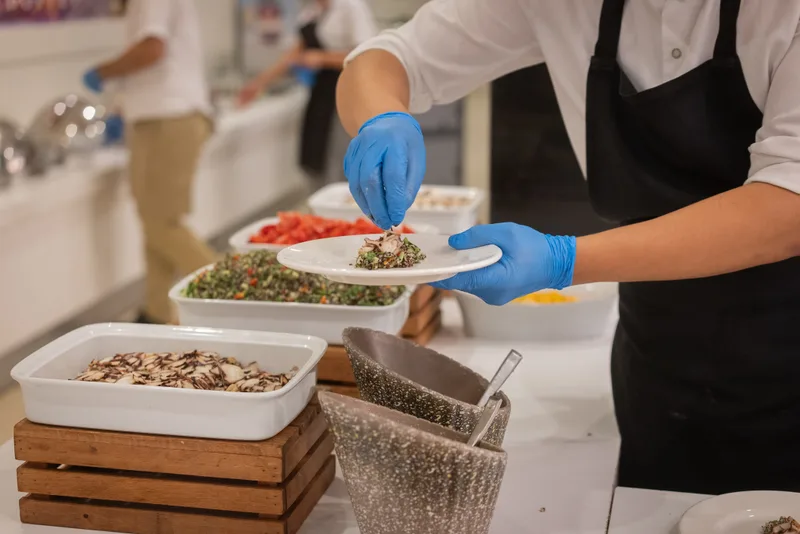 Catering staff in gloves portioning food from trays into serving dishes