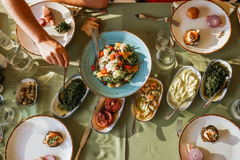 Overhead view of a table with a large salad bowl and shared plates of olives, shrimp, roasted tomatoes and greens