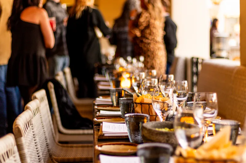 Long dinner table with glasses and printed menus, guests mingling behind