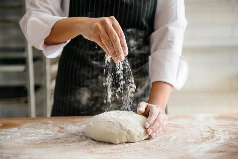 A baker in a striped apron dusting flour over a shaped round of dough on a flour-strewn bench
