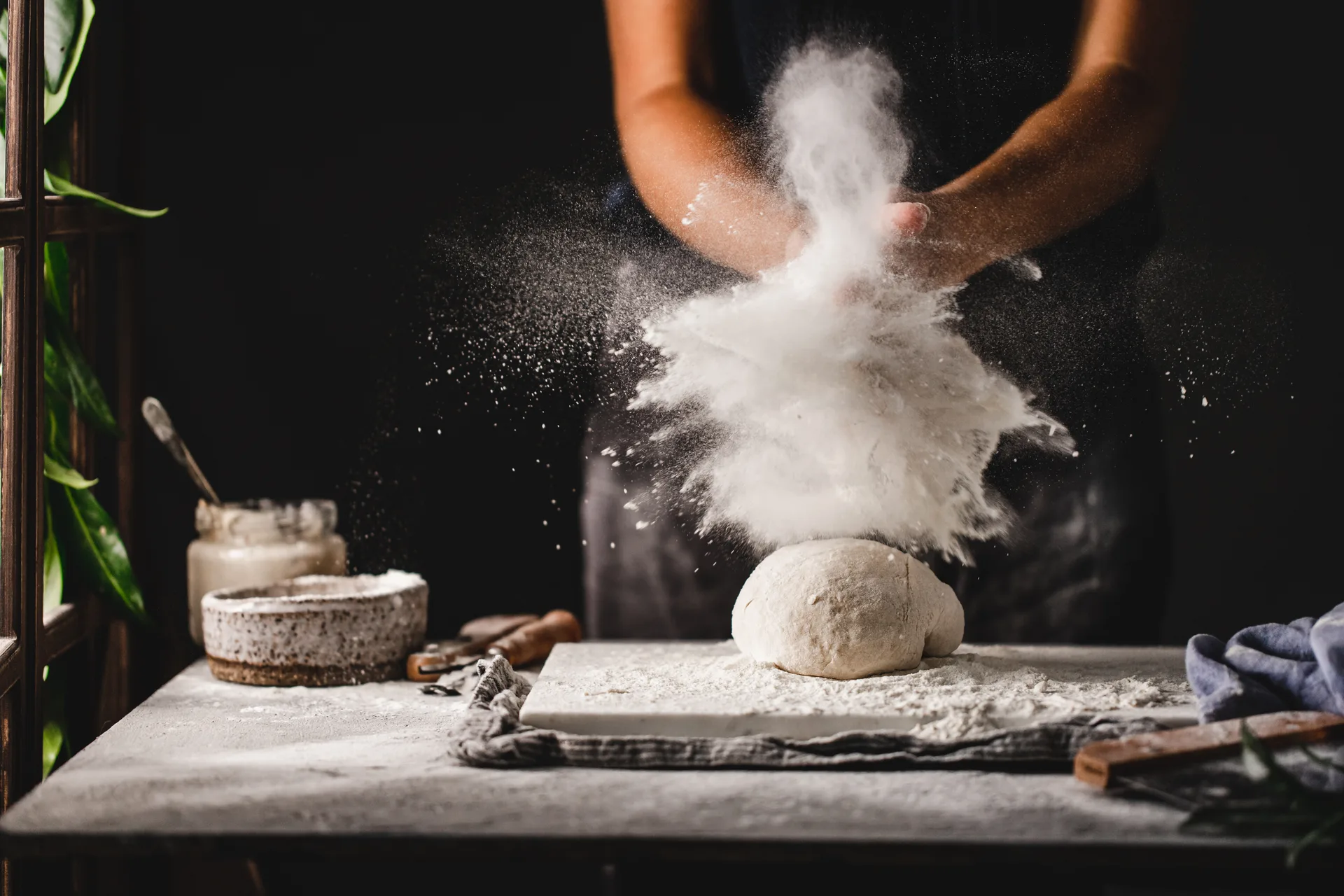 A baker clapping flour from their hands in a dramatic white cloud above a round of dough on a stone bench