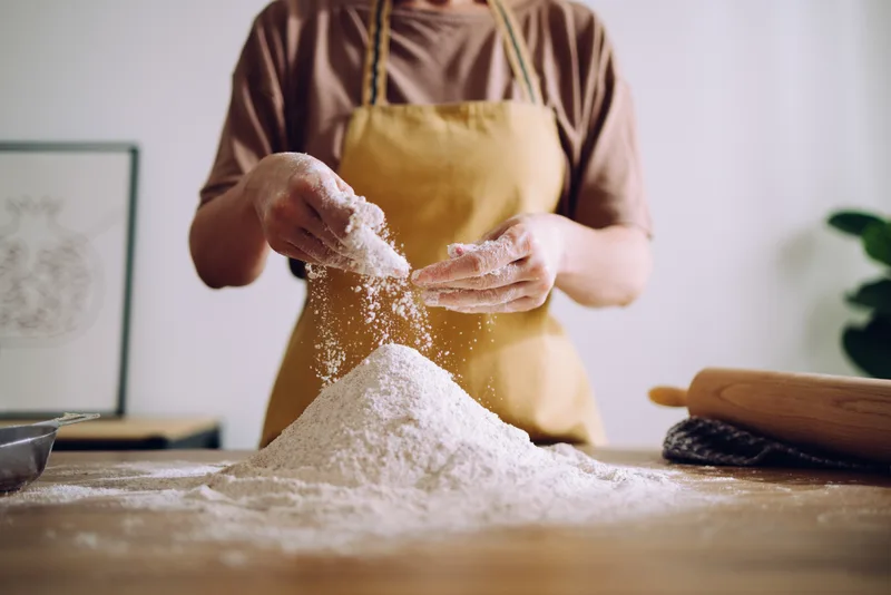 A workshop student in a yellow apron sifting flour onto a bench at the start of a sourdough class