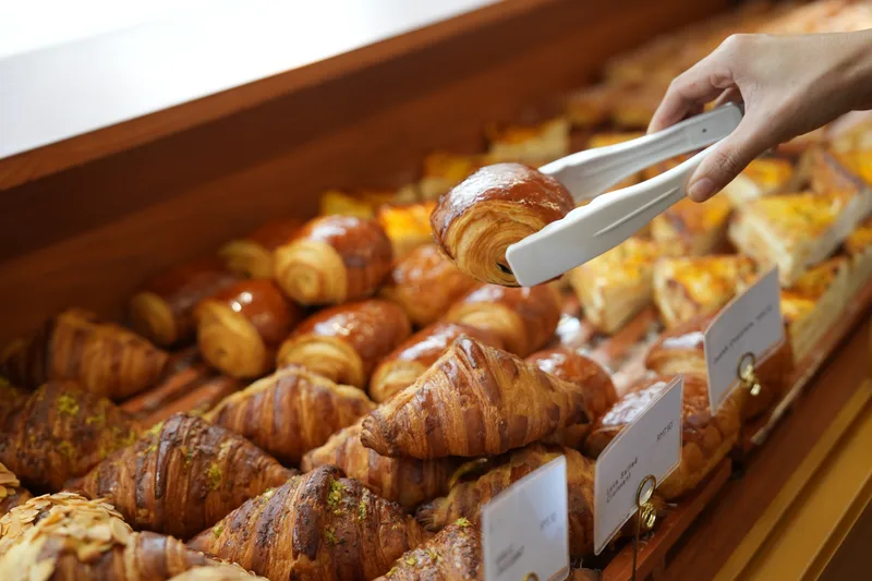 Tongs lifting a glazed pastry from The Corner's case, rows of croissants and morning buns with small paper labels below