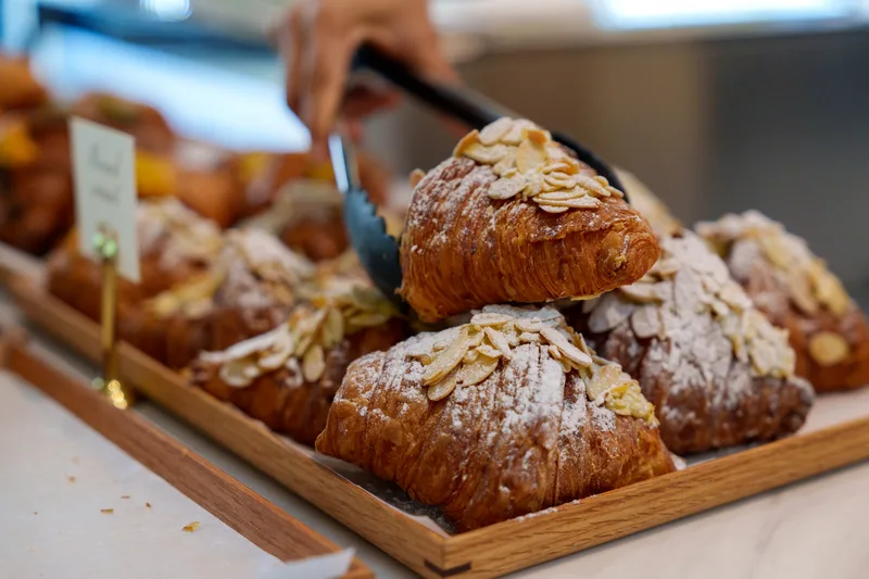 Sugar-dusted almond croissants lined up on wooden trays with tongs reaching for one