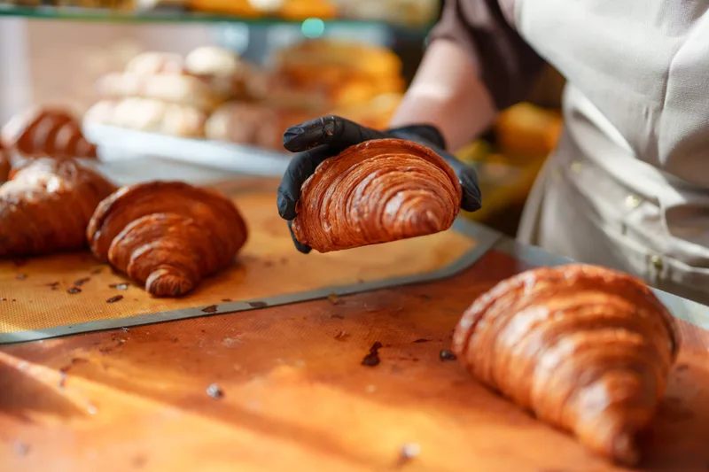 A baker in black gloves holding up a finished croissant above trays of fresh-baked croissants during a class