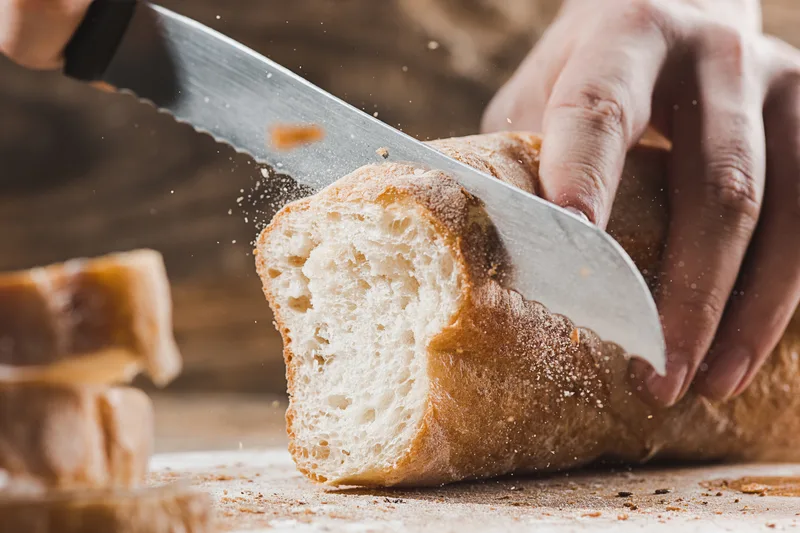 Hands slicing a fresh country loaf with a long bread knife, crumbs scattering across the board