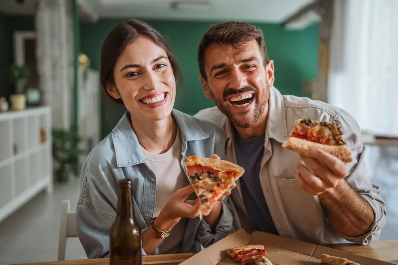 Two friends laughing on the couch, each holding up a slice of pizza