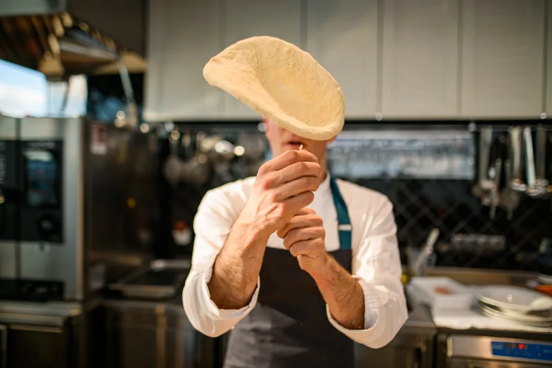 A pizza maker tossing a stretched round of dough into the air in the kitchen