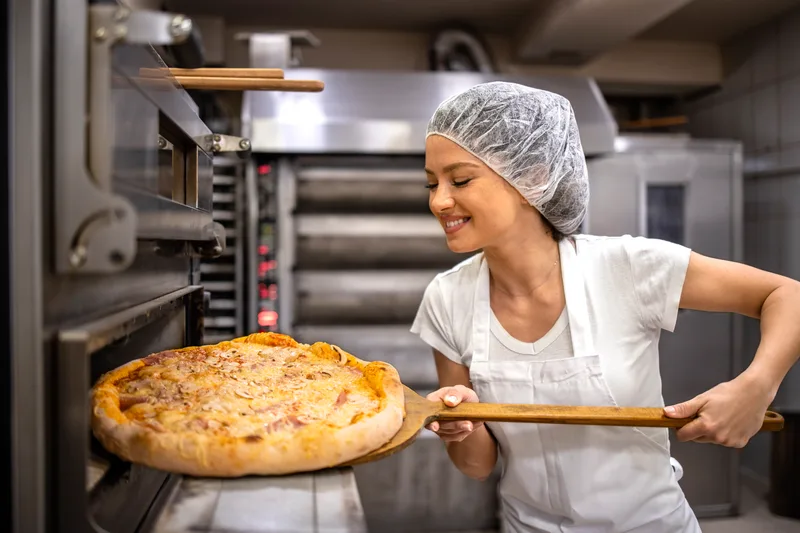 A smiling cook pulling a golden pizza out of the deck oven on a wooden peel