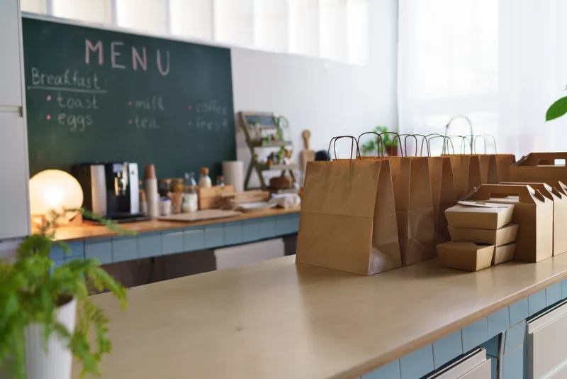 Paper takeout bags and boxes lined up on the pickup counter under a chalk menu board