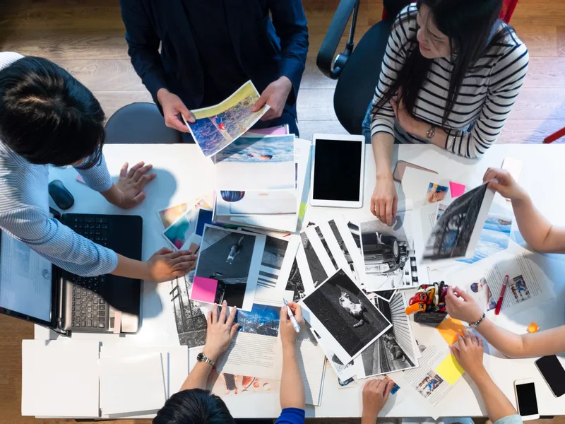 Overhead view of a design team spreading printed photographs and layouts across a white worktable, laptops open at the edges
