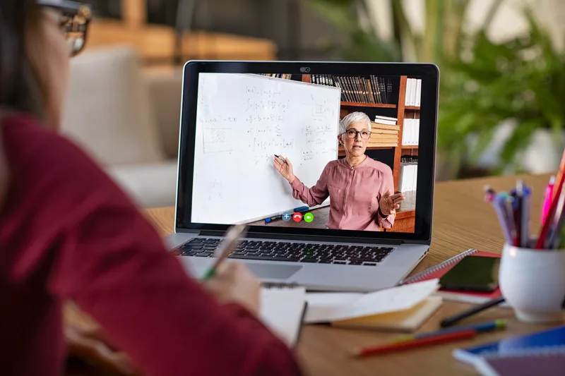 A team member takes notes while watching a colleague present at a whiteboard over a laptop video call