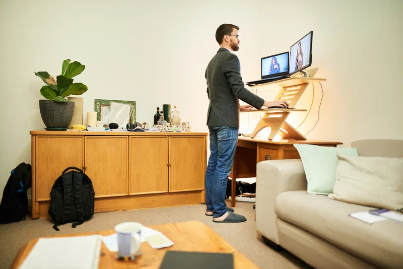 A remote worker stands at a home-office standing desk, joining a video call from his living room