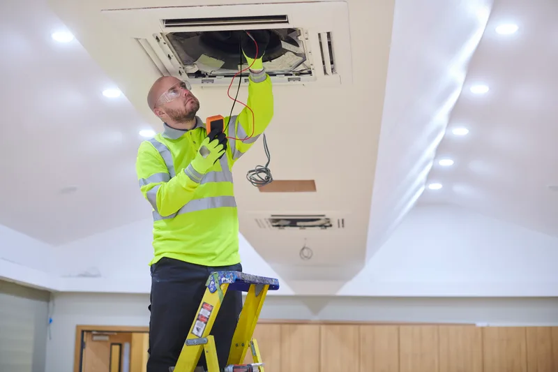 A technician in a hi-vis jacket stands on a ladder testing a ceiling-mounted air-conditioning cassette with a multimeter