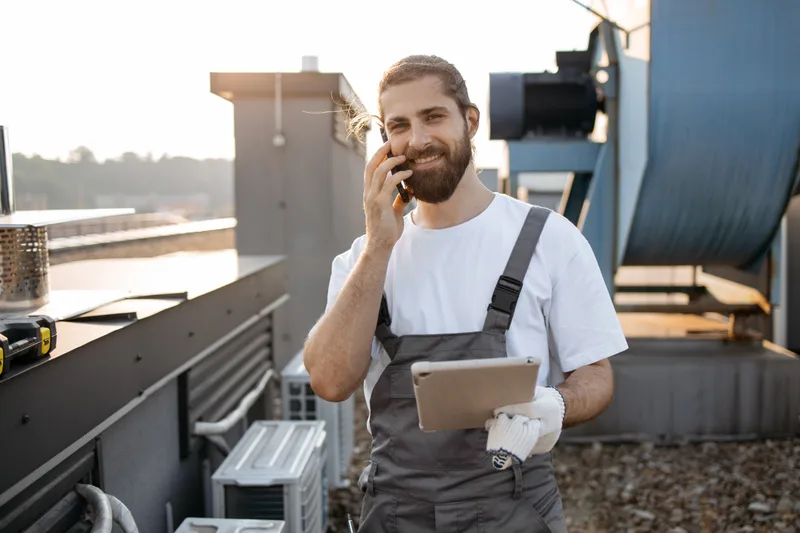 A smiling HVAC technician on a rooftop takes a customer call on his phone while holding a tablet with his day's jobs