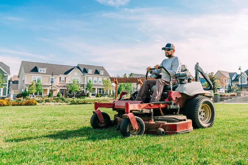 A lawn-care operator rides a zero-turn mower across a wide suburban lawn on a sunny day