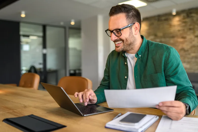 A smiling property manager in a green shirt reviews a printed report beside his laptop at a wooden office table